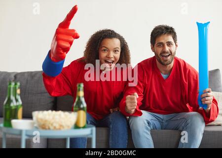 Portrait d'un couple de courses mixtes moderne regardant un match de sport à la télévision à la maison et applaudisse émotionnellement tout en portant des uniformes d'équipe rouge, espace de copie Banque D'Images