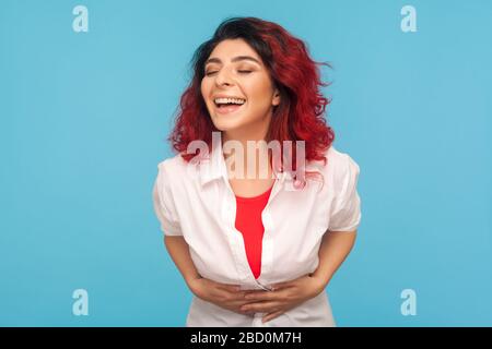 Rires, émotions positives. Portrait d'une femme heureuse avec des cheveux rouges fantaisie en chemise blanche tenant son ventre tout en riant fort après avoir entendu une blague. Banque D'Images