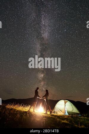 Romantique famille touristes ayant un repos au camping de nuit d'été dans les montagnes. Homme et femme debout à côté de feu de joie et de tente de glacer, tenant les mains sous le ciel étoilé plein d'étoiles et de voie lactée. Banque D'Images