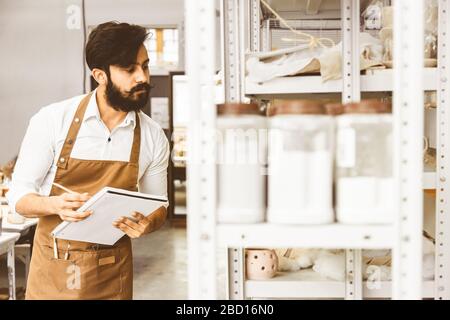 Jeune homme d'affaires séduisant un potter avec une barbe et une moustache travaille dans son atelier. Conserve les enregistrements et transcrites dans un ordinateur portable en inspectant Banque D'Images