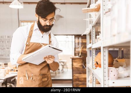 Jeune homme d'affaires séduisant un potter avec une barbe et une moustache travaille dans son atelier. Conserve les enregistrements et transcrites dans un ordinateur portable en inspectant Banque D'Images