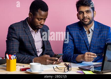 Tiré de deux hommes d'affaires portant des costumes assis au bureau, à l'aide d'un ordinateur portable et d'un téléphone portable. Homme indien partageant des idées avec un collègue afro-américain, Banque D'Images