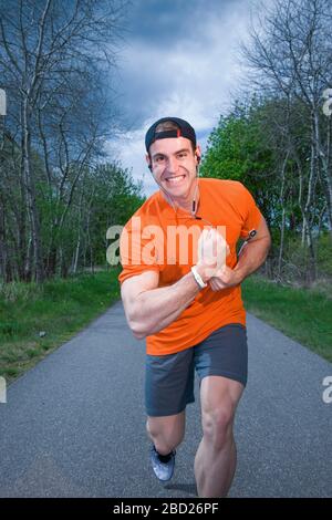 Vue frontale complète du corps d'un athlète de course dans une chemise orange dans une pose réussie sur un chemin pavé entre les arbres regardant la caméra. Banque D'Images