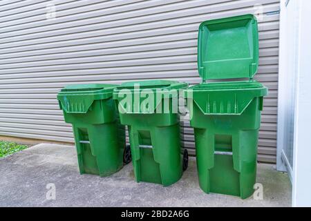 Une piqûre horizontale de trois poubelles en plastique vert s'aligne contre le mur extérieur d'un condominium. On a son couvercle vers le haut. Banque D'Images