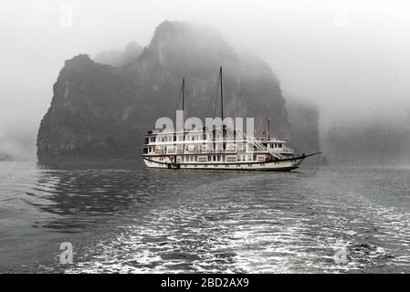 Photographie en noir et blanc d'un bateau touristique qui navigue devant l'une des nombreuses îles calcaires de la baie d'Halong par un jour brumeux - Vietnam, Asie Banque D'Images