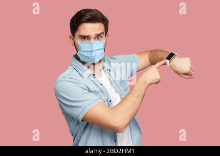 Le temps est écoulé. Portrait de jeune homme sérieux avec masque médical chirurgical en chemise bleue debout et regardant l'appareil photo, pointant sur sa montre intelligente. indoo Banque D'Images