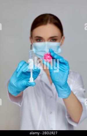 femme médecin en uniforme blanc, masque et gants en caoutchouc contenant seringue et modèle de virus sur fond gris. traitement en cas de pandémie. se concentrer sur la main Banque D'Images
