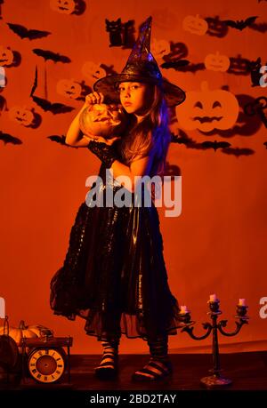 Petite sorcière portant un chapeau noir. Fête d'Halloween et décoration concept. Kid in en costume de sorcières épiques dans la lumière orange tient Jack o lanterne. Fille avec un visage sérieux sur fond orange avec décor Banque D'Images