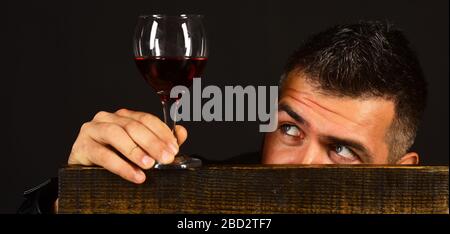 L'homme avec barbe tient un verre de vin rouge sur fond brun foncé. Degustator avec face cachée derrière une chaise en bois. Le sommelier a un goût de boisson chère. Concept de dégustation et de dégustation. Banque D'Images