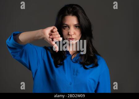 Femme adulte debout sur fond gris studio montrant le pouce vers le bas geste et exprimant le mécontentement à l'appareil photo. Portrait de la femme espagnole avec signe de Banque D'Images