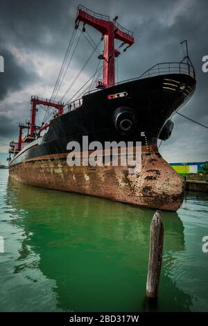 Vue grand angle d'un cargo noir et rouge rouillé et abîmé amarré à un quai et flottant sur une eau verte, sous un ciel nuageux menaçant Banque D'Images