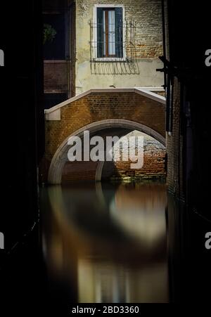 Photo nocturne d'un pont en pierre rond, partiellement caché et encadré par deux murs sombres, réfléchissant sur un canal vénitien Banque D'Images