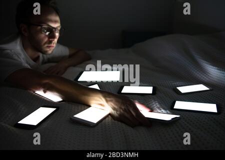 Dépendance aux gadgets, nomophobie, insomnie, troubles du sommeil. Ennuyer homme allongé dans le lit et prend l'un des nombreux smartphones actifs pour répondre au message sur social Banque D'Images