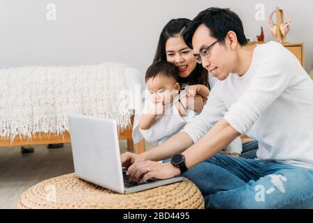 Père asiatique travaillant avec son ordinateur portable avec sa femme et son petit garçon asiatique mignon tout en étant assis au sol dans une chambre confortable à la maison. Banque D'Images