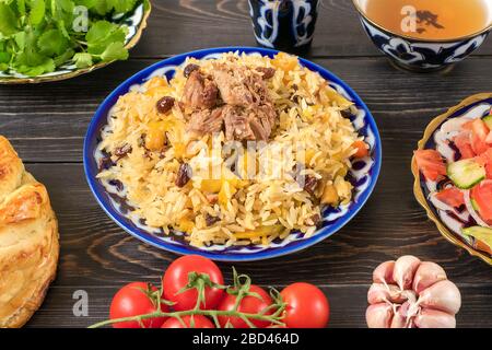 Pilaf ouzbek national avec viande, salade d'achichuk de tomate, concombre, oignon en plaque à motif traditionnel, coriandre, tomates cerises, pain garliche Banque D'Images