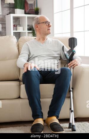 Un vieil homme solitaire assis sur le canapé dans une maison de soins qui tient des béquilles et regarde loin. Banque D'Images