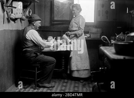 Femme qui verse une boisson pour un homme assis à une table dans une maison dans la Manche large, Queens CA. 1910-1915 Banque D'Images