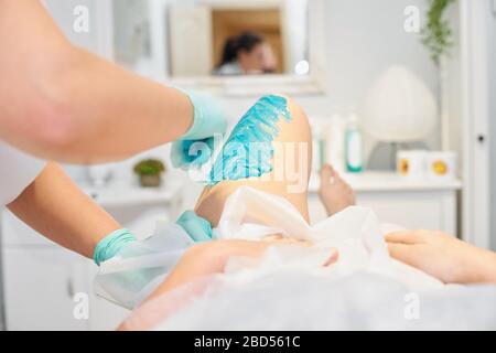 Concept d'épilation et de beauté -Beauticien dépirant les jambes des jeunes femmes avec du sucre liquide dans le centre thermal. Épilation des jambes avec des secoures turquoise Banque D'Images