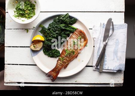 Une assiette avec truite cuite, kale et citron sur une table en bois blanc avec couverts et une serviette Banque D'Images