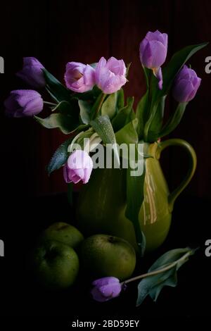 Un bouquet de tulipes lilas dans un pot vert avec des pommes vertes couvertes de gouttes de pluie sur fond noir Banque D'Images