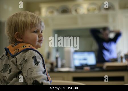 Enfant tout-petit assis sur la table à la maison pendant que maman travaille dans la cuisine pendant la période d'auto-isolation pendant la pandémie de coronavirus COVID-19 de 2020 Banque D'Images