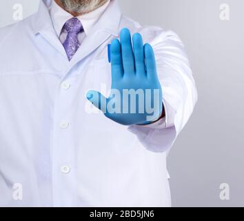 le médecin masculin dans un manteau blanc et des gants stériles bleus montre un geste d'arrêt avec sa main droite, fond blanc Banque D'Images