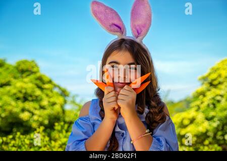 Portrait d'une jolie petite fille vêtue d'un lapin jouant avec des carottes, jardinage et s'amuser à l'extérieur en journée ensoleillée de printemps, célébration de Pâques Banque D'Images