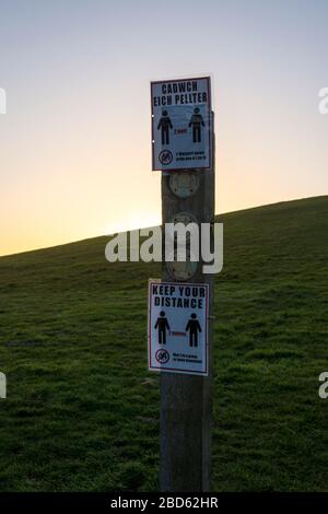 Un panneau en bois pour les marcheurs a des avis de distanciation sociale du coronavirus en anglais et gallois qui lui sont attachés. C'est le crépuscule avec un champ vide derrière. Banque D'Images