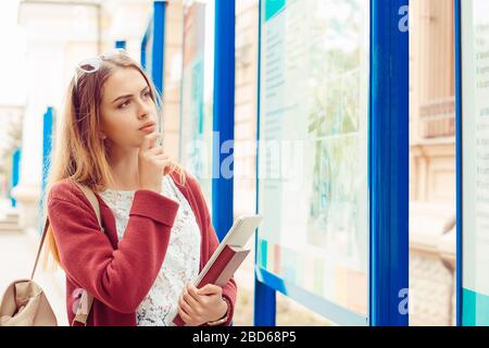 Fille confuse pensant à la carrière. Futur étudiant avec lunettes de soleil sac de livres dans la main regardant vers le haut des stands d'université pensant ce métier t Banque D'Images