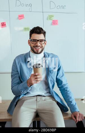 Casse maître regardant l'appareil photo et sourire avec tasse de café en papier près de la planche blanche avec feuille de calcul avec faire, faire lettrage et autocollants Banque D'Images