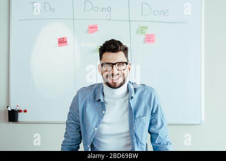 Scrum maître regardant l'appareil photo et sourire près de tableau blanc avec des autocollants et feuille de calcul avec à faire, faire, a fait lettrage Banque D'Images