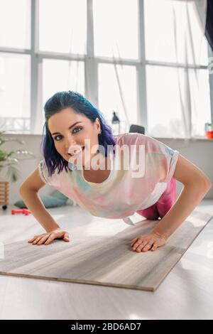 Fille avec des cheveux colorés souriant, regardant l'appareil photo et faire pousser sur le tapis de fitness dans le salon Banque D'Images