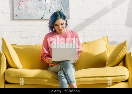 Freelance avec des cheveux colorés et un ordinateur portable souriant et assis sur un canapé jaune avec des jambes croisées dans le salon Banque D'Images