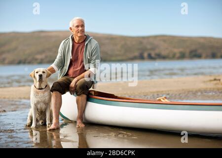 Heureux homme senior assis sur le bord d'un canoë avec son chien. Banque D'Images