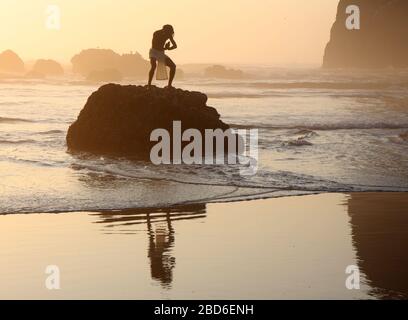 Un homme spirituel dans un zen pose au sommet d'un rocher sur une plage au coucher du soleil. Banque D'Images