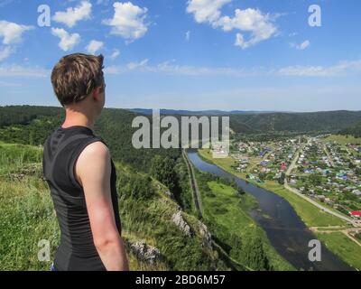 Un jeune homme au sommet d'une montagne regarde le paysage, les maisons de la vallée et le ciel bleu. Tourisme, voyage, escalade, victoire. Banque D'Images