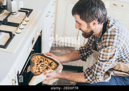 Jeune homme barbu prenant de la tarte aux baies en forme de coeur hors du four. Cuisine à la maison et sourire. Banque D'Images