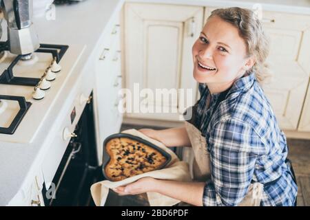 Jeune blonde femme prenant la tarte aux baies en forme de coeur hors du four. Cuisiner à la maison et sourire tout en regardant l'appareil photo. Banque D'Images