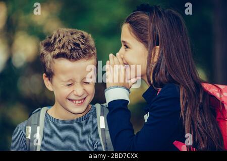 Fermez deux heureux amis avec des sacs à dos riant et s'amuser dans le parc. Une petite fille chuchote quelque chose dans une oreille de son ami avec le sourire an Banque D'Images