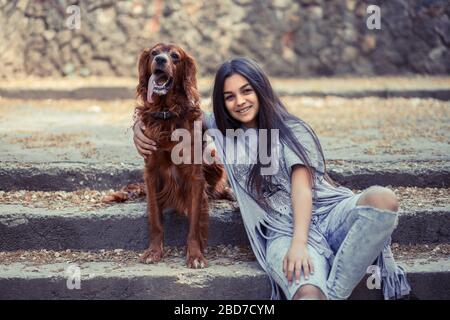 Jolie petite brunette embrassant avec un adorable chien assis sur des marches en pierre dans le parc et souriant avec plaisir à l'appareil photo. Banque D'Images