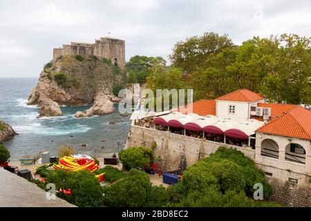 Vue sur les murs historiques de la vieille ville de Dubrovnik, dans le port ouest. La vue comprend le fort Lovrijenac, la plage et le restaurant de bord de mer. Banque D'Images