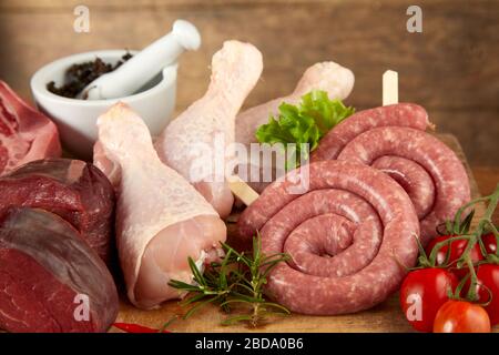 Assortiment de viandes fraîches brutes pour griller avec des pattes de poulet, des steaks de boeuf et des serpentins de saucisse sur un fond en bois avec des herbes et des épices en gros plan Banque D'Images