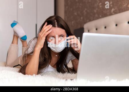 travail à distance. Une fille dans un masque de protection est allongée sur le lit, appelant sur son smartphone et regardant l'écran de l'ordinateur portable. Une personne malade travaille de hom Banque D'Images