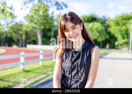 Heureuse belle jeune femme asiatique souriante à l'appareil photo pendant sa pause d'exercice le matin sur une piste de course avec des arbres flous et le ciel bac Banque D'Images