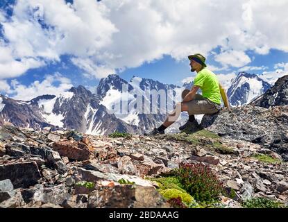 Randonneur avec chapeau et t-shirt vert assis sur la pierre et profiter de la vue des montagnes de neige au fond de ciel bleu Banque D'Images