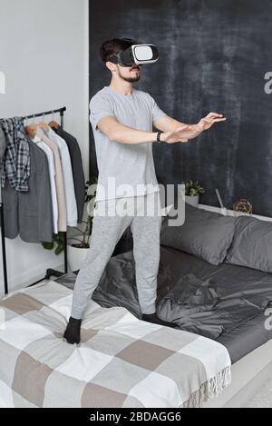 Jeune homme barbu dans la maison debout sur le lit et les mains gestuelles tout en jouant au jeu vidéo dans des lunettes de réalité virtuelle Banque D'Images