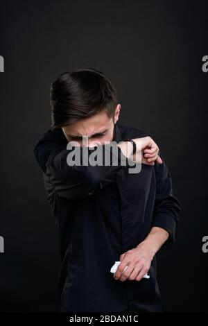 Jeune homme de brunette malade avec des symptômes de coronavirus tenant le napkin et toussant dans le coin du coude contre fond noir Banque D'Images