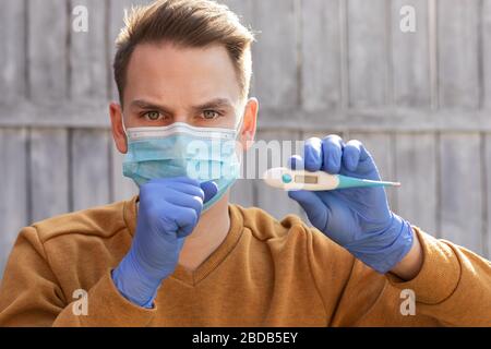 Le portrait d'un jeune homme contaminé par le covid-19 porte un masque et des gants, tenant un thermomètre numérique Banque D'Images