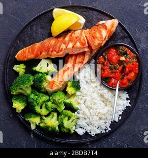 Steak de filet de saumon avec brocoli cuit à la vapeur, riz avec sauce tomate et épinards, quartiers de citron sur une plaque noire sur fond de béton foncé, couverts Banque D'Images