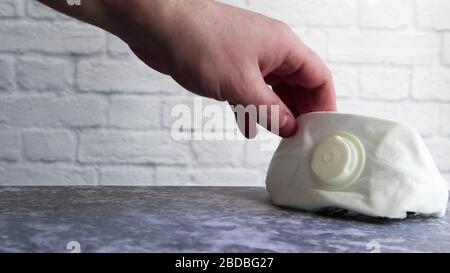 Une main masculine a levé ou met un masque médical blanc sur une table grise contre un mur de briques blanches avec espace de copie. Masques médicaux pendant les éclosions de di viral Banque D'Images